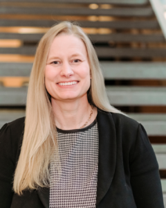 A woman with long blonde hair smiling and wearing a black blazer over a houndstooth blouse.