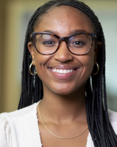 A woman with long dark braids wearing glasses and a white blouse, smiling.