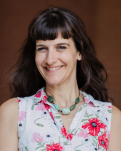 A woman with brown hair smiling, wearing a floral blouse and a green beaded necklace.