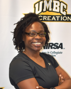 A woman with short brown curly hair standing in front of a University of Maryland, Baltimore County backdrop, wearing a black Under Armour T-shirt and glasses.