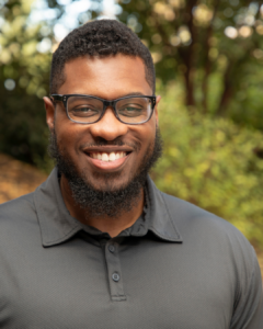 A man smiling, standing in front of greenery, wearing a dark gray polo shirt and glasses.