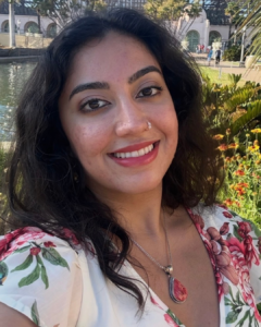 A woman smiles while standing outdoors, wearing a white floral top and a pink pendant necklace.